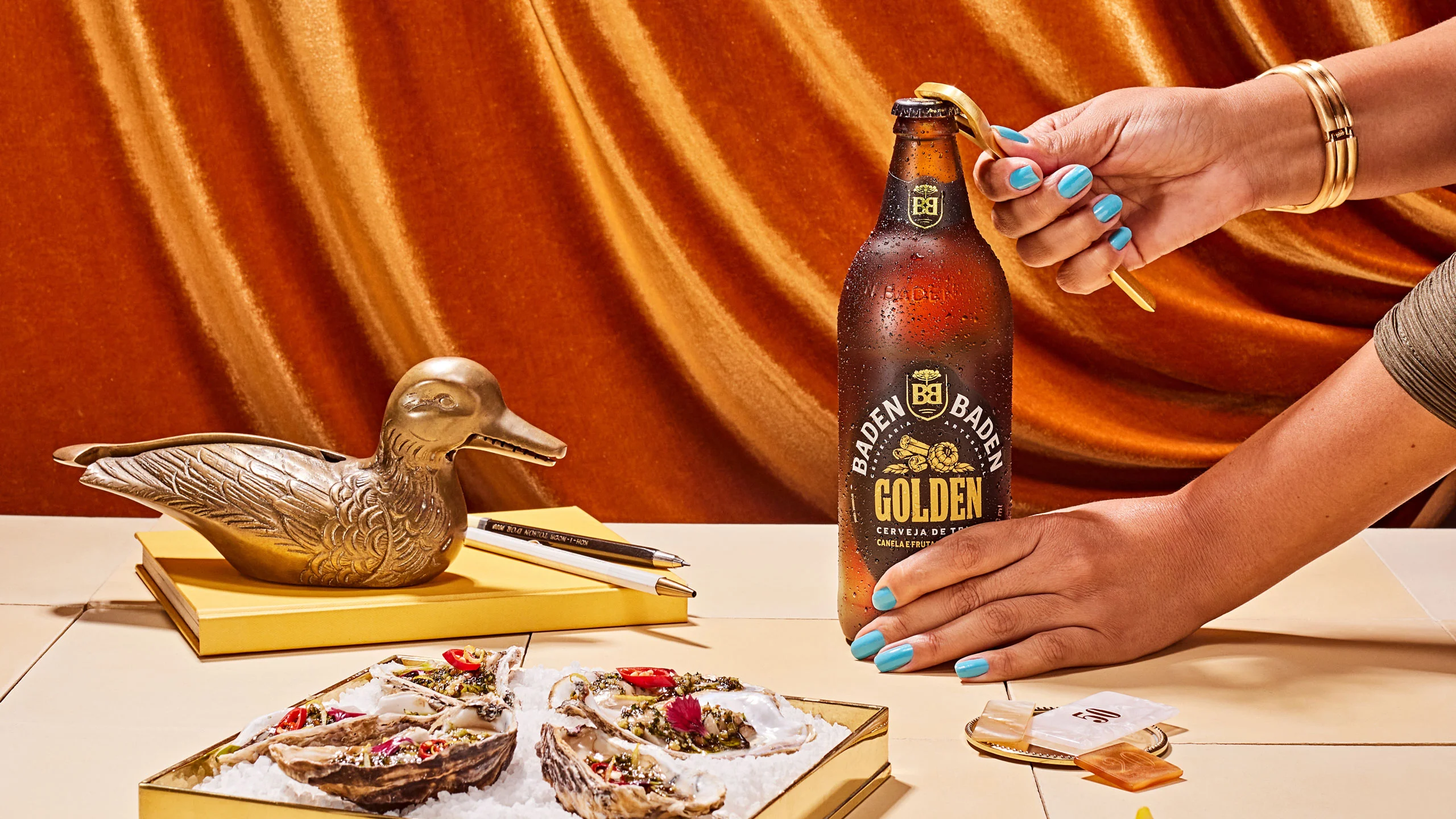 Mãos abrem cerveja Baden Baden Golden, com ostras frescas e pato de metal, em cenário laranja.